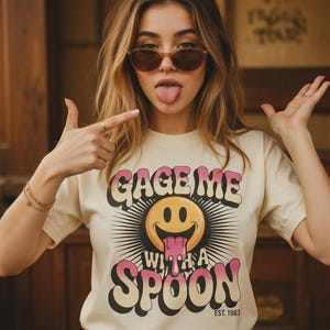 以下が含まれることがあります： ピンクと黒で「GAGE ME WITH A SPOON」の文字が入った、舌を出した笑顔のグラフィックが特徴のクリーム色のTシャツ。「EST. 1983」も下部にプリントされています。