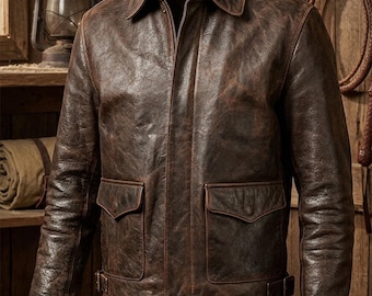 May include: A brown leather jacket with a distressed finish. The jacket features a collared neckline, a front zipper, two front pockets, and a belt detail at the waist. The jacket is hanging on a wooden wall.