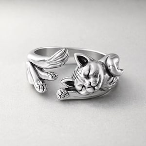 May include: A silver-toned open ring featuring a sculpted cat design. The cat is depicted in a curled, sleeping position with its head resting on one paw and its tail wrapping around the opposite side of the band, showing detailed fur texture.