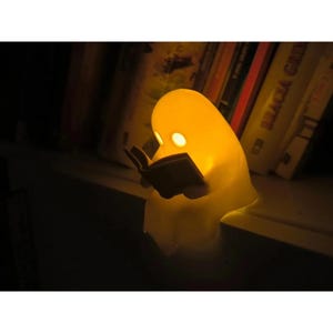 Puede incluir: Una pequeña lámpara con forma de fantasma de color blanco roto que descansa sobre un estante. La lámpara emite una luz amarilla cálida y presenta dos aberturas circulares para los ojos. La figura sostiene un pequeño libro abierto.