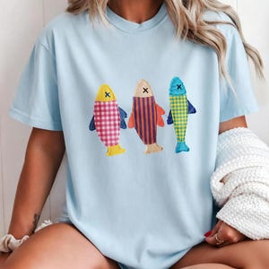 May include: A light blue short-sleeved t-shirt featuring a graphic of three stylised fish. The left fish has a pink and white gingham pattern, the middle fish has orange and purple vertical stripes, and the right fish has a yellow and green check.