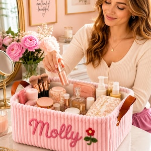 May include: A rectangular light pink corduroy storage basket with brown leather handles. The front features the embroidered name Molly in cursive pink script next to a small embroidered red and green flower. The basket is filled with various cosmetics.