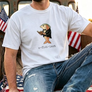 May include: A white short-sleeved t-shirt featuring a graphic of a fish wearing a black tuxedo and bowtie while reading a book. Below the fish, the text reads o-fish-iant in a black, typewriter-style font. The shirt has a crew neckline.