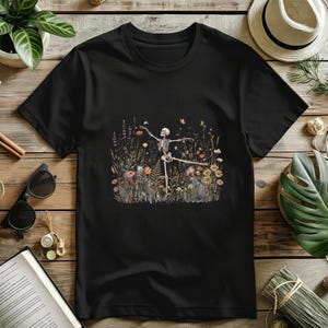 Könnte beinhalten: Ein schwarzes Kurzarm-T-Shirt mit einem zentralen Grafikdruck eines tanzenden menschlichen Skeletts, umgeben von einer detaillierten Illustration verschiedener Wildblumen, hoher Gräser und kleiner Schmetterlinge in gedeckten Erdtönen.