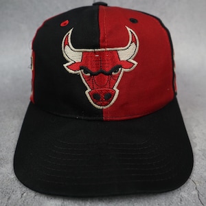 Könnte beinhalten: Eine Baseballkappe mit geteiltem Design, bei dem die linke Seite schwarz und die rechte Seite rot ist. Auf der Vorderseite befindet sich ein gesticktes Chicago Bulls-Logo, das einen rot-weißen Bullenkopf zeigt. Die Kappe hat einen schwarzen Schirm.