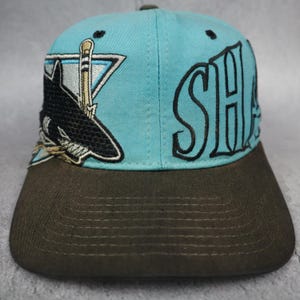 Pode incluir: Um boné de beisebol azul claro com aba marrom escura. A frente apresenta um gráfico bordado de um tubarão preto mordendo um taco de hóquei dentro de um triângulo, com o texto bordado em preto SHARKS no lado direito da copa.