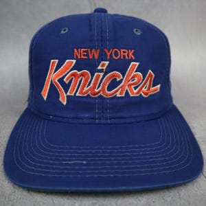 Op de afbeelding: Een koningsblauwe honkbalpet met de tekst NEW YORK in oranje borduursel boven het woord Knicks in een groter oranje scriptlettertype met een witte omlijning. De pet heeft een gestructureerde kroon en een gebogen klep met zichtbare stiksels.