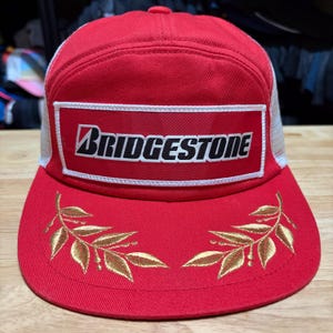Könnte beinhalten: Rote Trucker-Cap mit weißem Mesh-Rücken und einem rechteckigen Aufnäher mit dem "Bridgestone"-Logo in Schwarz und Weiß. Der Schirm der Kappe hat goldene, gestickte Blattmotive.