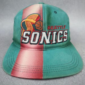 Puede incluir: Una gorra de béisbol color verde azulado con una franja roja vertical en el centro. La parte frontal muestra un logotipo bordado de los Seattle Sonics con un gráfico de baloncesto y el texto SEATTLE SONICS en hilo blanco, rojo y verde oscuro.
