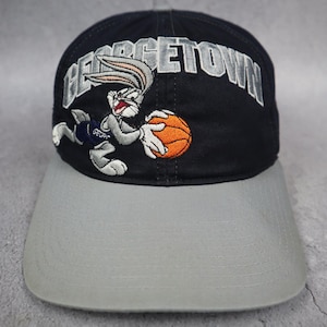 Op de afbeelding: Een zwarte honkbalpet met een grijze klep, voorzien van een geborduurde afbeelding van Bugs Bunny die een oranje basketbal vasthoudt. Het woord GEORGETOWN is in grijze blokletters boven het personage op de pet geborduurd.