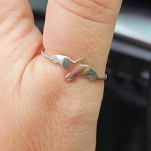 Puede incluir: Un anillo de plata con un diseño de un pequeño pájaro en vuelo en el pulgar, y un segundo anillo con una piedra negra larga y vertical en el dedo índice. Ambos anillos tienen bandas de plata pulida delgadas y se usan en los dedos.
