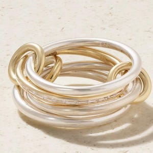 Puede incluir: Un anillo de varias bandas con aros entrelazados en tonos plata y oro. Una de las bandas plateadas está incrustada con una hilera de pequeñas piedras transparentes de corte baguette que captan la luz, creando una apariencia texturizada.
