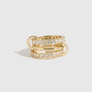 Puede incluir: Una pila de tres anillos dorados. Dos anillos están incrustados con piedras claras y brillantes, mientras que el anillo central es una banda lisa. Los anillos están dispuestos para crear un efecto de capas, mostrando un diseño de joyería moderno.