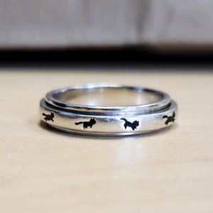 May include: A silver-colored ring with a spinning outer band. The band features several black cat silhouettes. The ring is set against a neutral background, highlighting the jewelry's design and craftsmanship. The ring is a piece of cat-themed jewelry.