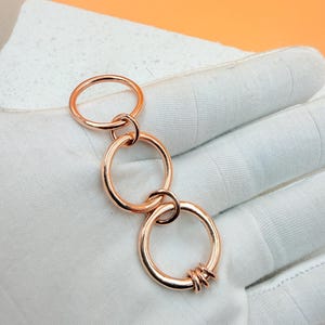 Op de afbeelding: Een roségouden sleutelhanger met drie onderling verbonden ringen. De onderste ring heeft verschillende kleinere ringen. De sleutelhanger wordt getoond op een wit oppervlak met een oranje achtergrond.