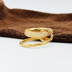 Op de afbeelding: Een gouden ring met een uniek ontwerp. De ring heeft een band bedekt met kleine, sprankelende stenen en een andere gladde, gouden band die eromheen wikkelt. De ring staat tegen een bruine achtergrond.