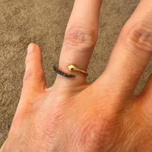 May include: A gold-toned open ring featuring a snake head design on one end and a tapered tail end encrusted with small black stones. The band is adjustable, wrapping partially around the finger with a polished finish on the snake head.