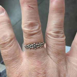 Può includere: Un anello color argento caratterizzato da una fascia continua di piccoli fiori in rilievo. Il metallo presenta una finitura leggermente ossidata nelle zone incavate tra i petali, che mette in risalto il motivo floreale attorno al dito.