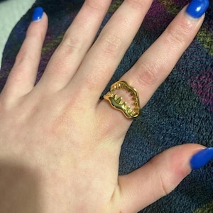 May include: A gold-toned ring featuring a stylized open mouth design with visible pointed teeth. The ring is crafted with a polished metallic finish and has an open-ended structure that wraps around the finger, showcasing a distinct jaw-like shape.