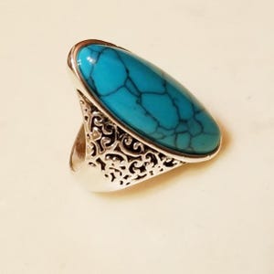 May include: A silver ring with a large, oval turquoise stone with dark veining. The ring has an ornate, openwork design on the band, adding a decorative element to the jewellery.