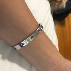 May include: A silver cuff bracelet with star cutouts. The bracelet is worn on a person's arm. The bracelet is made of a shiny, reflective metal. The stars are evenly spaced around the bracelet.