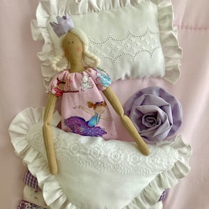 Puede incluir: Muñeca hecha a mano con cabello rubio y corona morada, vestida con un vestido rosa con diseño de bailarina. Descansa sobre una cama de almohadas blancas con bordes con volantes y una rosa de tela morada. La ropa de cama es rosa.