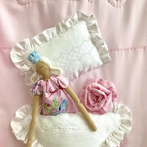 Puede incluir: Una muñeca de tela rosa con un vestido de hada y una corona azul, colocada sobre una almohada blanca con borde con volantes. También se ven una rosa rosa y otra almohada blanca. El fondo es una tela rosa claro con detalles bordados.