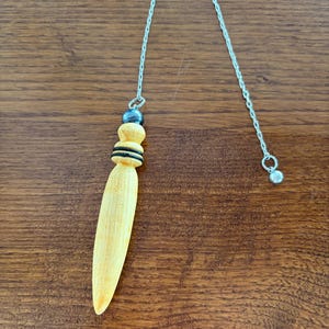 Puede incluir: Un péndulo de madera con una cadena plateada. El péndulo tiene un cuerpo en forma de lágrima de color amarillo claro, con anillos negros y amarillos cerca de la parte superior. Una cuenta gris oscuro y una cadena plateada completan el diseño. El péndulo está sobre una superficie de madera marrón.