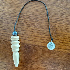 May include: A wooden pendulum with a light finish, featuring a carved design and a pointed end. It is suspended by a black cord, with a small silver charm depicting a lotus flower. A small, dark gray bead is attached to the cord.