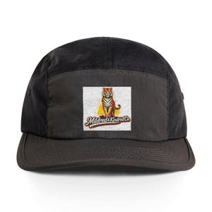Puede incluir: Gorra negra de cinco paneles con un parche cuadrado con una ilustración de un tigre. El tigre es naranja y negro, sentado sobre un fondo amarillo. El texto "Mildred & Kindred Co." está debajo.