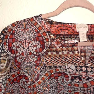 May include: A patterned blouse with a mosaic design in red, orange, and brown. The blouse has a V-neck and short sleeves. The tag reads "Chico's" and size "2". The blouse is hanging on a pink hanger. The pattern is intricate.