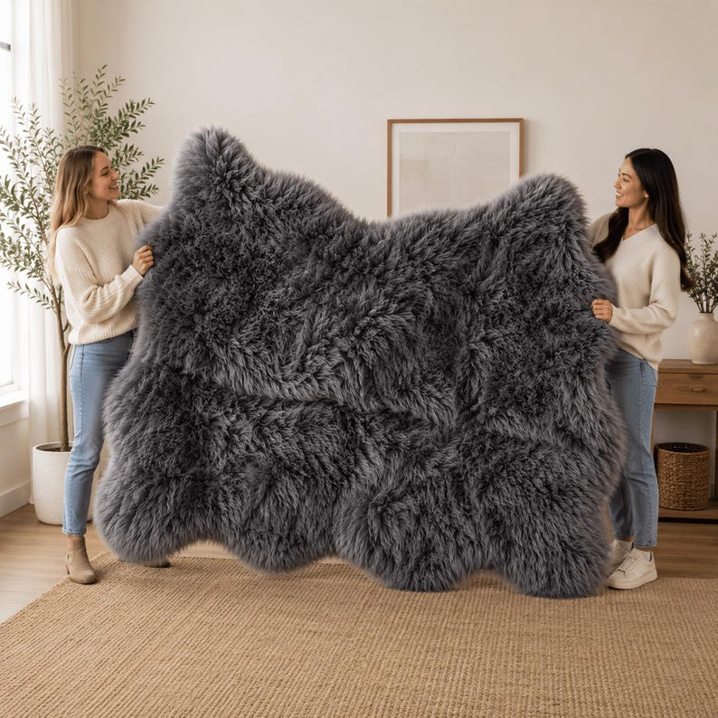 May include: A large, rectangular, dark gray sheepskin rug with a thick, long-pile texture. The rug features an irregular, natural edge shape and a dense, fluffy appearance, showcasing a deep charcoal color throughout its entire surface area.