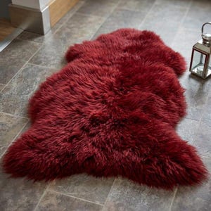May include: A deep red, plush faux fur rug with an irregular, organic shape and long, soft fibers. The rug features a dense, shaggy texture that covers the entire surface, providing a thick and layered appearance across its length and width.