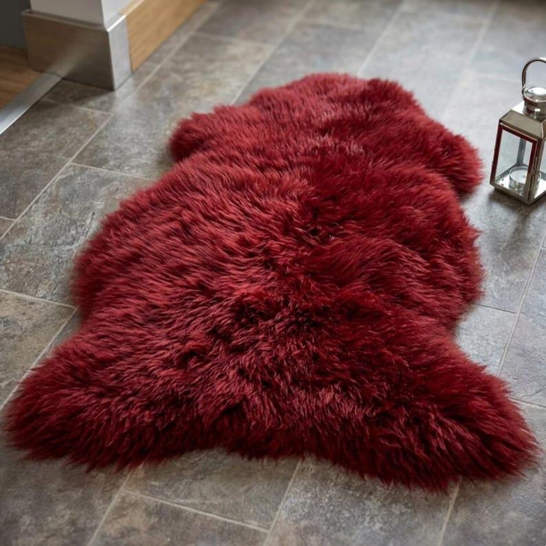 May include: A deep red, plush faux fur rug with an irregular, organic shape and long, soft fibers. The rug features a dense, shaggy texture that covers the entire surface, providing a thick and layered appearance across its length and width.