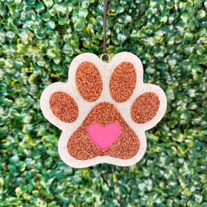 May include: A hanging paw print ornament featuring a white glittery base with copper-colored glitter pads and a central pink heart. The ornament is suspended by a thin black cord, showcasing a textured, sparkling finish on all its components.