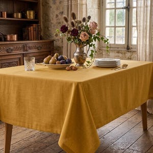 May include: A mustard yellow linen tablecloth covers a wooden table. A silver vase holds a bouquet of roses and other flowers. A plate of fruit, a stack of plates, and a glass are on the table. The table is in a room with a wooden floor and a stone wall.