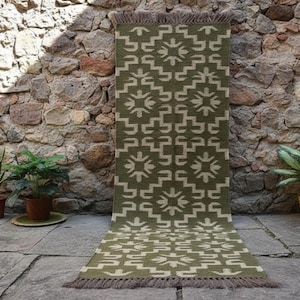 Puede incluir: Una alfombra rectangular verde oliva con un patrón geométrico en blanco roto. La alfombra tiene un borde con flecos y se muestra contra una pared de piedra. La alfombra mide aproximadamente 152 cm de largo.