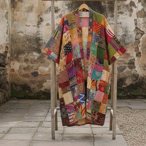 Puede incluir: Una bata tipo kimono de patchwork con una vibrante mezcla de colores y patrones. La bata está hecha de varios cuadrados de tela cosidos, creando un diseño único y artístico. Cuelga de un estante de madera contra una pared de piedra texturizada.
