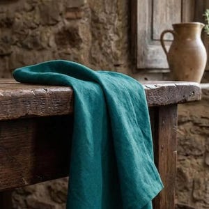 May include: A teal-colored linen table runner draped over a rustic wooden table. The fabric has a soft, flowing texture. The background features a stone wall and a wooden window frame, creating a warm, inviting atmosphere. A ceramic pitcher is visible.