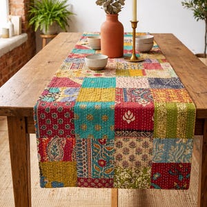 May include: A vibrant patchwork table runner in a range of colours, including red, blue, yellow, and green. The runner is on a wooden table, with a terracotta vase, bowls, and a candle holder. The table is approximately 180 cm long.