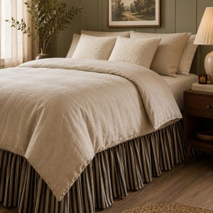 May include: A bed dressed in a solid beige duvet cover and matching pillowcases, featuring two textured cream-coloured accent pillows. The bed is finished with a pleated bed skirt showcasing a vertical black and beige striped pattern.