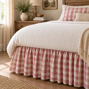 Puede incluir: Un faldón de cama con un patrón de cuadros rojos y blancos y un diseño con volantes. La cama está vestida con una funda nórdica de color blanco roto, una funda de almohada a cuadros rojos y blancos, un cojín lumbar y una manta color crema.