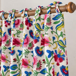 May include: A white fabric curtain featuring a vibrant floral pattern with red, pink, blue, and yellow flowers and green vines. The curtain is suspended from a wooden rod by several fabric ties knotted at the top, creating a gathered drape.