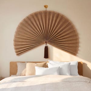 Könnte beinhalten: Eine große, fächerförmige Wanddekoration aus hellbraunem Holz hängt über einem Bett mit weißer Bettwäsche. Der Fächer hat eine Quaste und ist an einer beigefarbenen Wand befestigt. Das Bett hat ein hölzernes Kopfteil und mehrere Kissen.