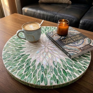 May include: A round, handmade mother-of-pearl tray with a green leaf design. A blue mug with coffee, a lit candle, and a book are on the tray. The tray is on a wooden table.
