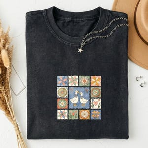 Op de afbeelding: Een zwart t-shirt met ronde hals en een centrale afbeelding van twee witte eenden, omringd door een raster van twaalf kleurrijke vierkante tegels met bloemen-, ster- en hartpatronen. Een zilverkleurige ketting met een sterretje ligt op de kraag.