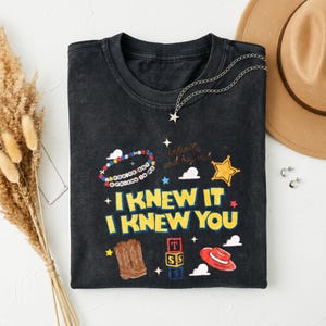 May include: A black crew neck t-shirt featuring a graphic print with the text I KNEW IT I KNEW YOU in yellow letters. The design includes illustrations of cowboy boots, a red hat, a yellow star, alphabet blocks, and a beaded bracelet.