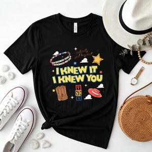 May include: A black short-sleeve t-shirt featuring a graphic print with the text I KNEW IT I KNEW YOU, a beaded bracelet, a yellow star, brown cowboy boots, a stack of alphabet blocks, and a red cowboy hat with white cloud shapes.