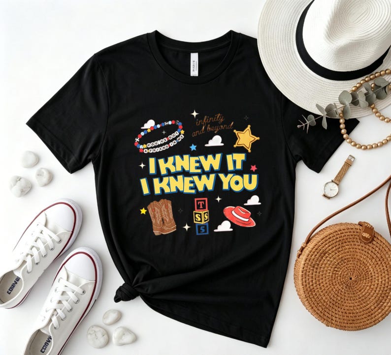 May include: A black short-sleeve t-shirt featuring a graphic print with the text I KNEW IT I KNEW YOU, a beaded bracelet, a yellow star, brown cowboy boots, a stack of alphabet blocks, and a red cowboy hat with white cloud shapes.