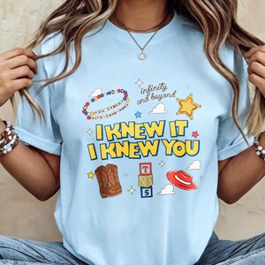 May include: A light blue t-shirt featuring a graphic print with the text "I KNEW IT I KNEW YOU", "infinity and beyond", and "YOU'VE GOT A FRIEND IN ME". The design includes illustrations of a beaded bracelet, a star, cowboy boots, blocks, and a hat.